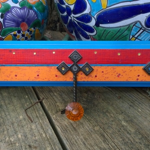 May include: A wooden wall-mounted coat rack with three hooks. The rack is painted in bright blue, orange, and red stripes. Each hook has a decorative metal cross and a colorful glass knob.