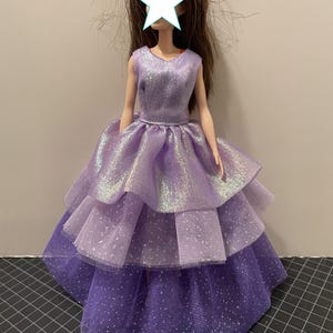 May include: A doll wearing a layered purple dress. The dress has a sleeveless bodice and a tiered skirt with a shimmering, iridescent fabric. The skirt has a dotted pattern on the bottom layer.