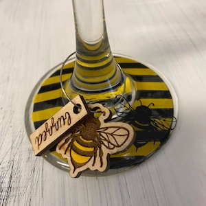 May include: A wine glass charm with a black and yellow striped base and a wooden bee charm with the word "Gurly" written on it.