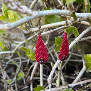 Könnte beinhalten: Ein Paar handgefertigte rote Makramee-Ohrringe in Tropfenform. Jeder Ohrring hat eine kleine, dunkelrote Perle am unteren Ende und antike bronzefarbene Ohrhaken. Die Ohrringe werden vor dem Hintergrund grüner Blätter und Zweige präsentiert.