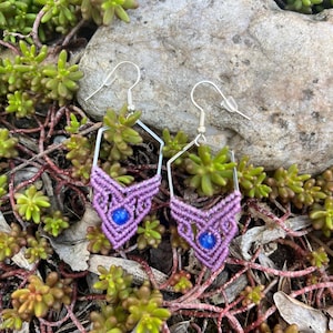 May include: Purple and blue macrame earrings with a geometric design. The earrings are hanging from silver hooks.