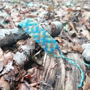 Pode incluir: Uma pulseira da amizade tecida com um padrão de cestaria repetitivo em tons de turquesa brilhante, verde claro e castanho suave. A pulseira tem dois fios finos trançados que se estendem de uma extremidade, feitos de linha de cor a condizer.