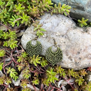 May include: Olive green macrame earrings with a detailed woven pattern. The earrings, teardrop-shaped, hang from bronze-coloured ear wires. The earrings are shown on a rock, surrounded by green and red-tinged succulents.
