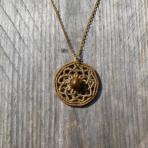 Op de afbeelding: Een gouden ketting met een ronde hanger met een bruine steen omgeven door een delicaat gouden draadontwerp.