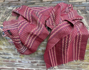 May include: A burgundy scarf with a diamond pattern and vertical stripes. The scarf has a fringed edge with blue, yellow, and green threads. The scarf is made of woven fabric.