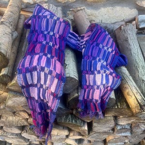 Könnte beinhalten: Zwei gestrickte Schals in den Farben Lila, Rosa und Blau mit einem strukturierten Muster liegen auf einem Holzstapel.