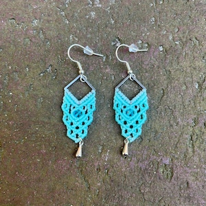 May include: A pair of turquoise macrame earrings with silver-coloured metal accents. The earrings have a geometric design with a small silver teardrop charm at the bottom. The earrings are displayed on a textured, earthy-toned surface.