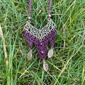 Può includere: Una collana in macramè viola e oro con un ciondolo in metallo decorativo e ciondoli a forma di foglie.