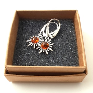 May include: A pair of silver earrings with amber sun-shaped charms. The earrings are in a brown box with a black foam interior.