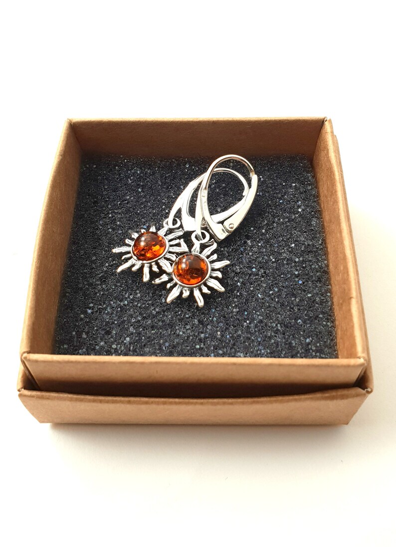 May include: A pair of silver earrings with amber sun-shaped charms. The earrings are in a brown box with a black foam interior.