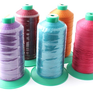 Puede incluir: Cinco carretes de hilo en diferentes colores: morado, granate, azul verdoso, naranja y rosa. Los carretes están sobre bases verdes.