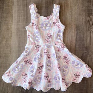 Puede incluir: Un vestido sin mangas con un estampado de Minnie Mouse y Daisy Duck. El vestido tiene un dobladillo festoneado y está decorado con rayas rosas, moradas y blancas. El vestido está hecho de una tela suave y ligera.