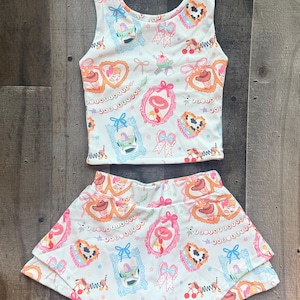 May include: A white two-piece outfit comprising a tank top and a skirt. The set is decorated with a colourful pattern of framed characters, bows, and hearts. The tank top has a scoop neck, and the skirt has a layered design.