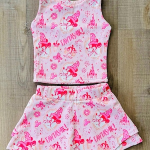 May include: Pink two-piece outfit featuring a tank top and skirt. The set is patterned with a repeating design of Mickey Mouse in a sorcerer's hat, castles, and the word "FANTASTIC!". The skirt has a layered, flared design.
