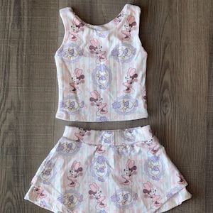 Può includere: Un completo a due pezzi composto da una canotta e una gonna. Il set è decorato con un motivo ripetuto di Minnie Mouse e altri personaggi in una combinazione di colori rosa chiaro, viola e bianco. La gonna ha un design a strati.