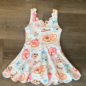 May include: White sleeveless dress with a scalloped neckline and hem. The dress features a repeating pattern of framed cartoon characters, hearts, bows, and other decorative elements in pink, blue, and orange.