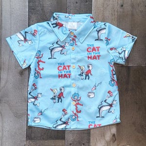 May include: A light blue short-sleeved button-down shirt with a pattern of "The Cat in the Hat" characters. The shirt has a yellow button closure and a collar.