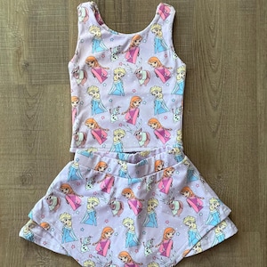 May include: A two-piece children's outfit featuring a light purple tank top and skirt. The set is covered in a repeating pattern of cartoon characters, stars, and reindeer. The skirt has a layered design.