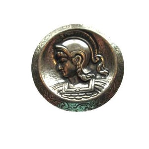 May include: A round, silver-toned button with a detailed relief of a classical figure wearing a helmet. The figure is in profile, set within a decorative border. The button has an antique appearance.