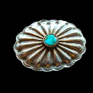 May include: An oval-shaped silver concho pin featuring a scalloped edge and a series of radiating embossed lines. A small, round, bright turquoise stone is set securely in the center of the metallic piece, creating a distinct contrast.