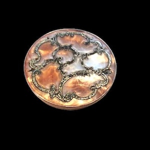 May include: A round, decorative button with a swirling, ornate design. The button features a mix of colors, including brown, white, and gold, with a metallic border. The button is set against a black background.