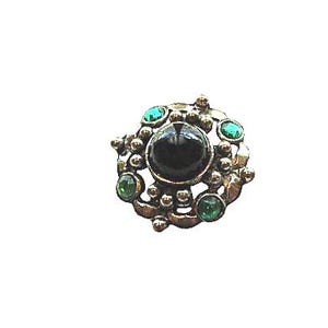 May include: A circular brooch featuring a large, smooth, black central cabochon stone surrounded by a metallic frame with small silver-toned spheres and four smaller, round, vibrant green stones set at intervals around the perimeter.
