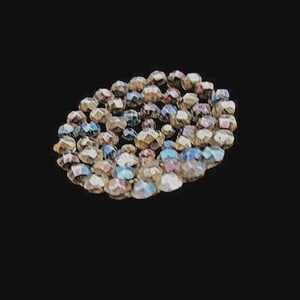 May include: A circular arrangement of faceted glass beads featuring a metallic, iridescent finish with shifting hues of bronze, silver, and blue. The beads are tightly clustered together, creating a textured, reflective surface with a multi-faceted appearance.