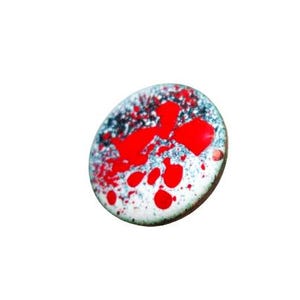 May include: A small, oval-shaped pendant with a white base, speckled with blue and red paint. The red paint forms irregular shapes and splatters across the surface. The pendant has a dark brown edge.