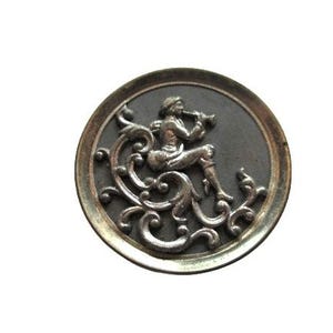 May include: A round, antique silver-toned button with a detailed relief design. The central image depicts a seated figure playing a wind instrument, surrounded by ornate scrollwork. The button has a dark gray background and a silver border.