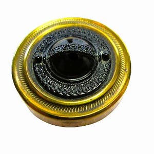 May include: A round, antique button with a gold-colored metal frame. The button features a central black oval surrounded by a detailed, dark gray design. The outer rim has a textured, ridged pattern.