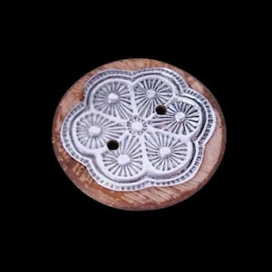 May include: A round wooden button with a silver floral design. The silver design is in the shape of a flower with two holes in the center. The button is a light brown color.
