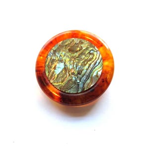 May include: A round, decorative button with a tortoiseshell-like outer ring. The center features a shimmering, iridescent abalone shell design. The button is set against a white background, highlighting its colors and textures.