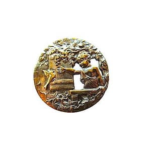 May include: A circular metallic button featuring a relief design of a figure reaching toward a bird perched on a structure. The scene is framed by intricate foliage and branches, rendered in a polished gold and dark bronze tone finish.