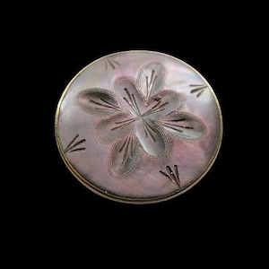 May include: A circular mother-of-pearl button featuring an iridescent, light purple surface with a carved, six-petaled flower design at the center. The button has a smooth, polished finish and is framed by a thin, metallic gold-toned edge.