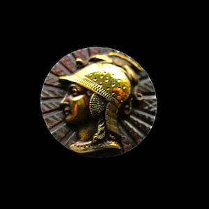 May include: A circular metallic button featuring a relief profile of a person wearing a detailed helmet. The design includes a sunburst pattern radiating from the central figure, with a polished gold-tone finish on the helmet and dark accents.