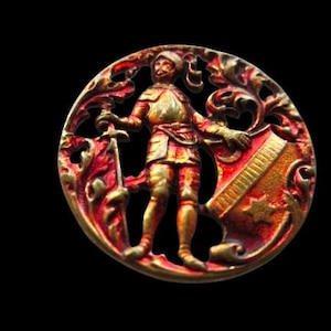 May include: Ornate, circular button with a detailed relief of a medieval knight. The knight is depicted in gold armor, holding a sword and shield, set against a red and gold filigree background. The button has a vintage, antique appearance.