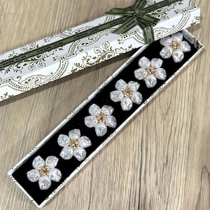 May include: A gift box with a white and gold paisley pattern, containing six white flower-shaped ornaments with gold centres. The box is open, revealing the ornaments on a black velvet base. A green ribbon with the words "BEST WISHES" is tied around the box.