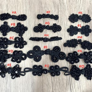 May include: A collection of fifteen black decorative frog fasteners labeled A through O, featuring intricate cord work, bead embellishments, and knotted designs. These fasteners vary in shape and size, showcasing diverse patterns of loops and textures.