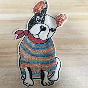 May include: A white dog wearing a blue, pink and silver sequined sweater with a red bandana around its neck. The dog has black eyes and a black nose. The dog is sitting on a light brown wooden surface.