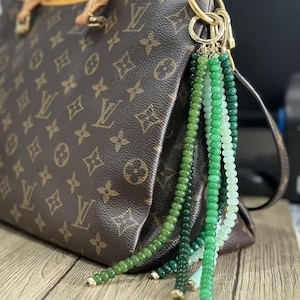 May include: Brown designer handbag with a gold chain and a green beaded tassel. The handbag features a repeating pattern of interlocking letters and floral designs. The tassel is made of various shades of green beads.
