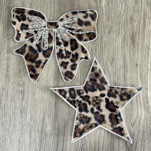 Puede incluir: Un lazo y una estrella con estampado de leopardo con detalles de pedrería. El lazo tiene una cadena plateada y un centro de pedrería. La estrella tiene un borde blanco y está cubierta de pedrería.