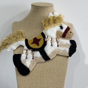 May include: A decorative, handmade horse applique with a white body, black hooves, and a tan mane and tail. The horse has a black and gold saddle with a red star design. The applique is displayed on a tan mannequin.
