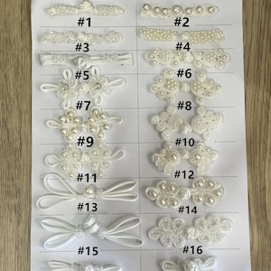 May include: A collection of eighteen white decorative frog buttons arranged on a white sheet, numbered one through eighteen. Each button features intricate cord work, some accented with small white beads or pearls in various symmetrical patterns.