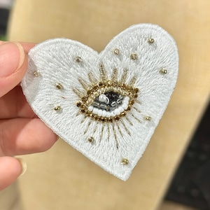 Op de afbeelding: Wit, hartvormig geborduurd embleem met een oogontwerp. Het oog is gedetailleerd met gouden en zwarte kralen, omringd door gouden stralen. Kleine gouden kralen zijn verspreid over de witte stof. Geschikt voor kleding of accessoires.