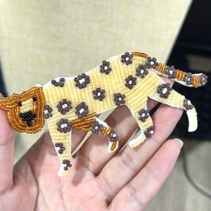 Puede incluir: Broche de leopardo con cuentas. El broche presenta un cuerpo amarillo con manchas marrones en forma de flor. La cabeza y la cola están detalladas con cuentas marrones. El leopardo está en posición de caminar.