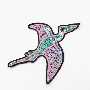 May include: A pink and green iridescent sequined iron-on patch of a flying dinosaur.