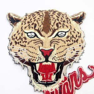 May include: Embroidered iron-on patch featuring a snarling leopard head with brown, white, and red details. The patch has a red embroidered word "Wild" below the leopard head.