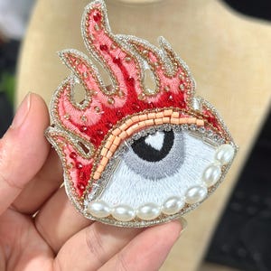May include: Embroidered eye brooch with red flame design. The eye features a white iris, a black pupil, and a row of pearls along the bottom. The flames are outlined in gold and accented with red beads.