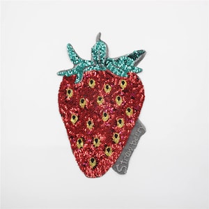 Peut inclure: Un patch de fraise en sequins rouge et vert avec le mot "Strawberry" brodé en bas.