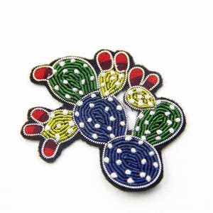 Puede incluir: Un parche de cactus bordado de colores con hilo verde, azul, amarillo y rojo. El cactus está hecho de varias secciones y tiene cuentas blancas para los detalles.